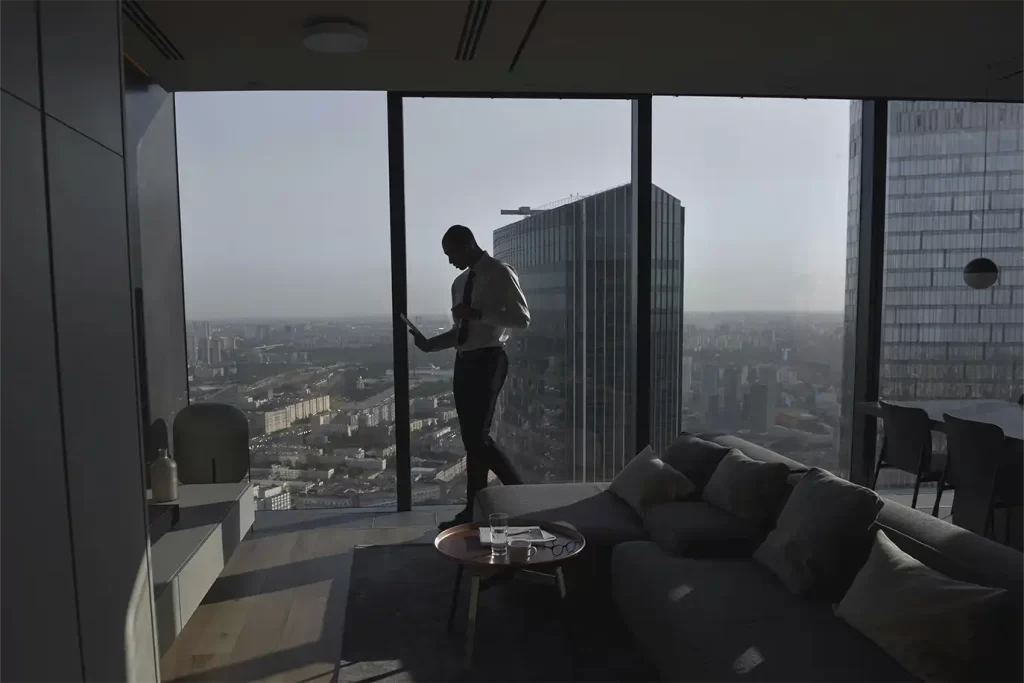 Ejecutivo trabajando con tablet frente a un ventanal de gran altura con vistas panorámicas a la ciudad en suite moderna.