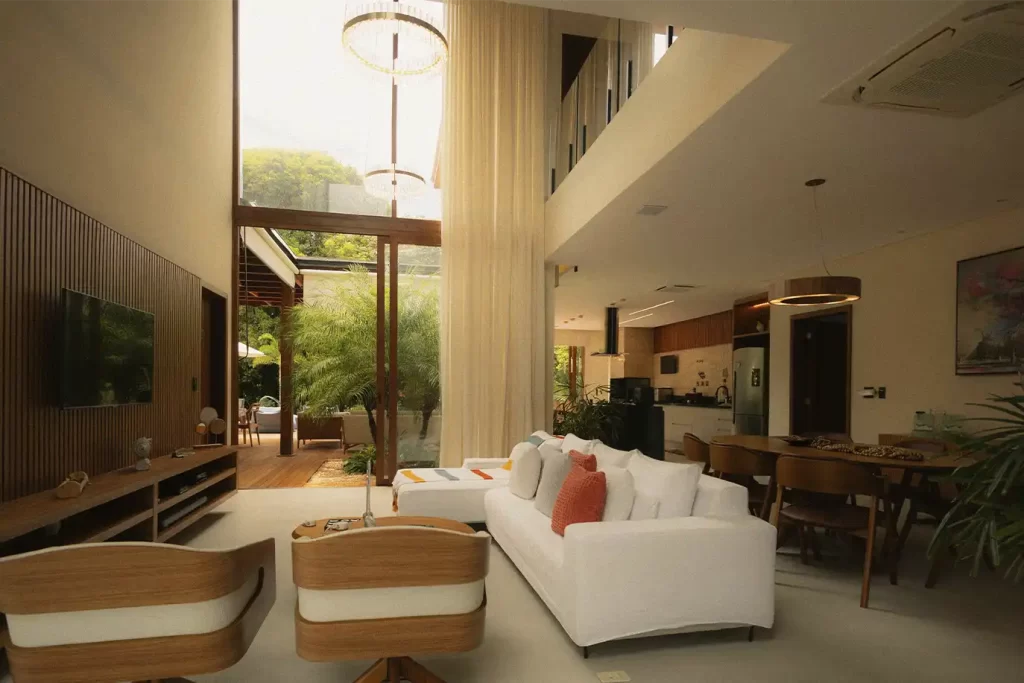 Sala de suite de lujo con techos de doble altura, sofá blanco, comedor de madera y ventanal hacia jardín privado iluminado.