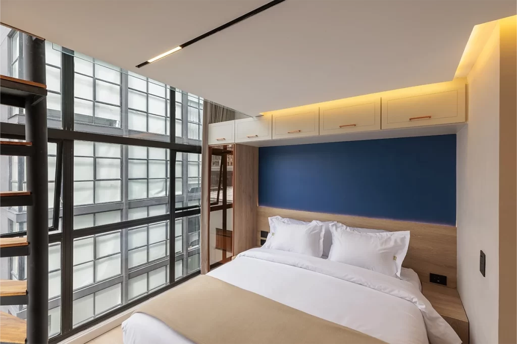 Dormitorio de loft con cama doble, cabecero azul y ventanal de doble altura en Bogotá.